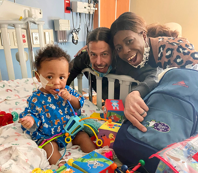 A mom and dad smiling with their child who is in a hospital bed with toys and a bag.