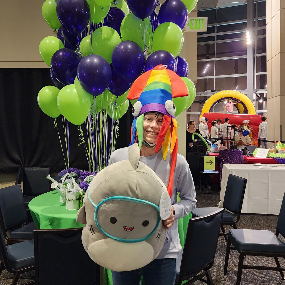 Adlyn, a teenage oncology patient, with the rainbow squid hat on holding a large stuffed cartoon character in front of green and purple balloons and a bounce house in the distance.