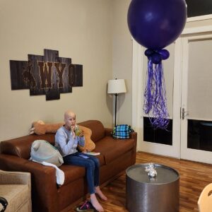 Adlyn, a teenage oncology patient, sitting on a brown couch in her apartment with a bundle of the purple balloons from the event floating above the small round table in front of her.