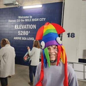 Adlyn, a teenage oncology patient, with a rainbow squid hat on in front of a blue wall that says, Welcome to Denver, CO the Mile High City. Elevation 5280 feet above sea level.