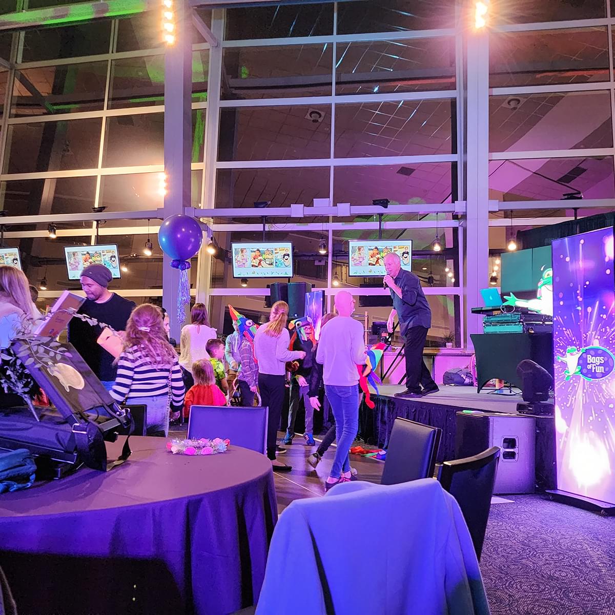 A man on stage in an indoor space signing while adults and kids, including Adlyn, a teenage oncology patient, are dancing. There are purple lights and a digital screen with the Bags of Fun logo.