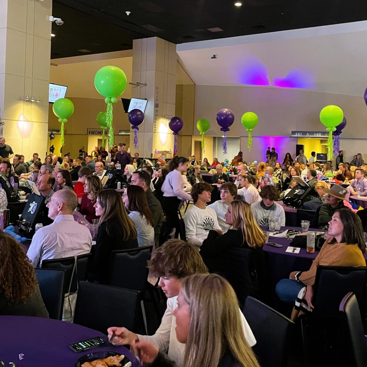 Lots of people sitting at round tables eating and talking at the event. There are green and purple balloons all around as well.
