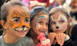 3 kids with face paint smiling at an event. One kid has tiger face paint, another a dolphin, and the third is a mouse.