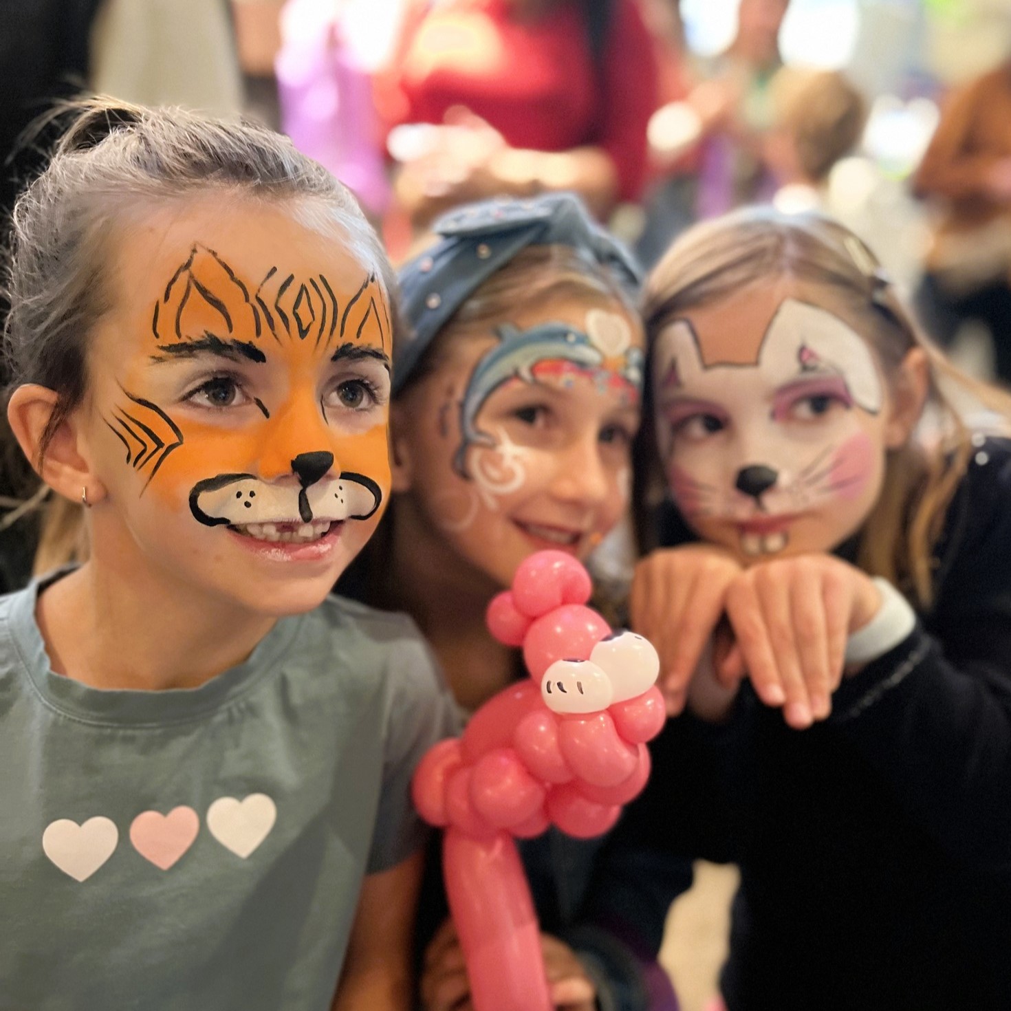 3 kids with face paint smiling at an event. One kid has tiger face paint, another a dolphin, and the third is a mouse.