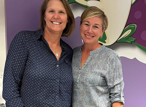 Susan Buckheit and Catherine Winter pose for a photo. There is a purple wall behind them with a large image of the Bags of Fun logo on it. You can only see part of the logo.