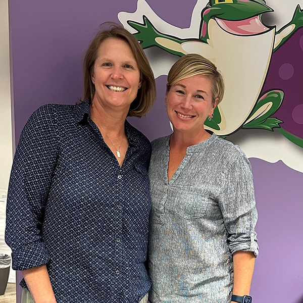 Susan Buckheit and Catherine Winter pose for a photo. There is a purple wall behind them with a large image of the Bags of Fun logo on it. You can only see part of the logo.