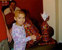Gabby in her pajamas smiling while unwrapping a present. There are a couple other presents on the table and there is a man in the background smiling.