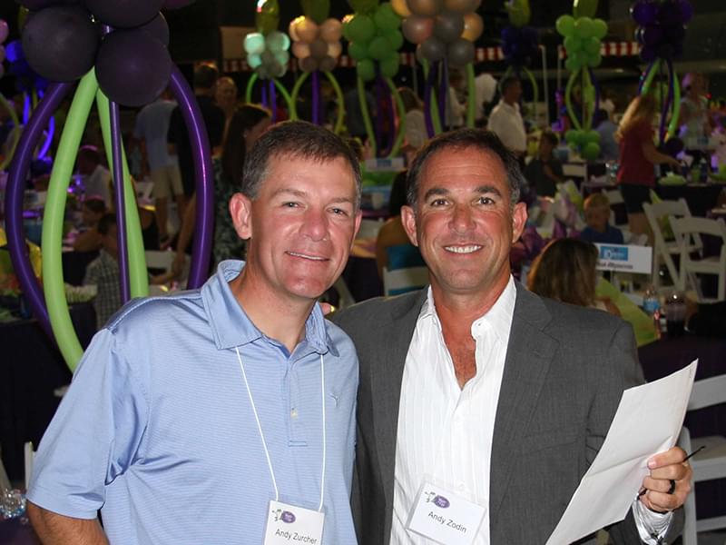 To men, both names Andy, posing for a photo at the Bring Out the Bags Event. We see lots of people in the background with green and purple balloon decorations.