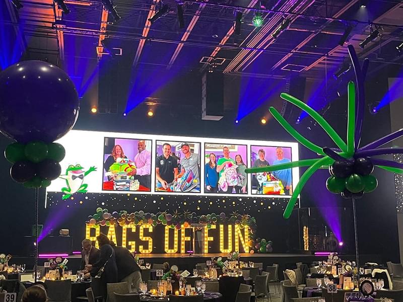 A stage with large light up yellow letters with the words Bags of Fun written out sitting on the stage. There is a large screen above the letters with images of volunteers packing and holding bags and there are tables in front of the stage nice set ready for guests to arrive. There are a couple people in front of one table getting ready to site down.