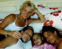 3 family members are lying down on a couch or bed with Gabby. Gabby is in the middle. The pillows they are lying on have lard red flower prints. They are relaxed and smiling.