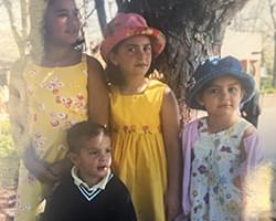 4 kids of different ages are all standing next to each other. 3 of them are looking off to the right but one, who is Gabby, is looking forward. Gabby has on a blue hat and a flower dress with a pink shall. Two of the older girls are wearing yellow summer dresses and one has a pink hat on. There is also a much younger boy wearing a dark color sweater with a white collared shirt underneath.