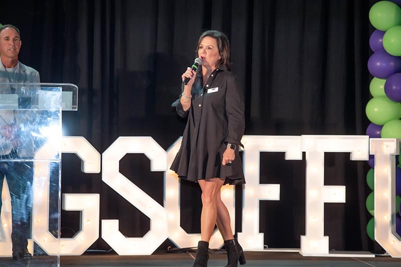 Tammy on stage wearing a black dress and black boots. She is speaking with a microphone in her hand and seems to be walking across the stage as she speaks. We can see large white letters with lights in them sitting on the stage behind her. We see the letters GSOFFU. It appears the words spell Bags of Fun but some letters are not in the image. There are also purple and green balloon on the right part of the stage behind the letters. There is also a man in a light blue blazer with a tshirt underneath and some dark slacks on. He is standing to the left of the stage behind a clear podium.