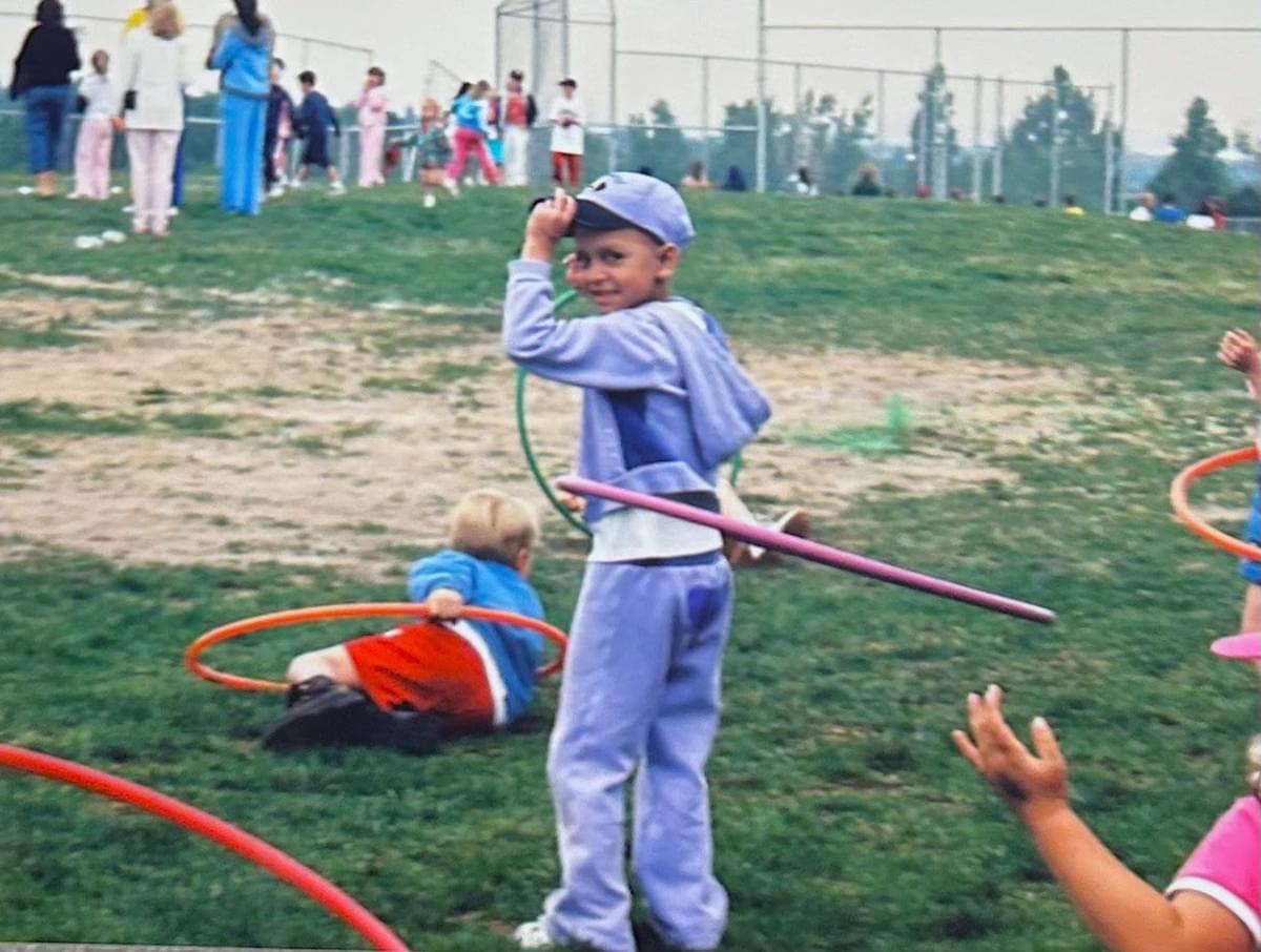 Gabby's in a purple jump suite and matching hat standing and doing the hula hoop. She is smiling, holding the brim of her hat and looking at the person holding the camera. She is one of the last ones with the hula hoop up. There are a couple hula hoops on the ground and one kid looks like he just fell on the ground with his hula hoop. They are standing in a patchy green and brown grassy area and we see lots of people standing in the background.
