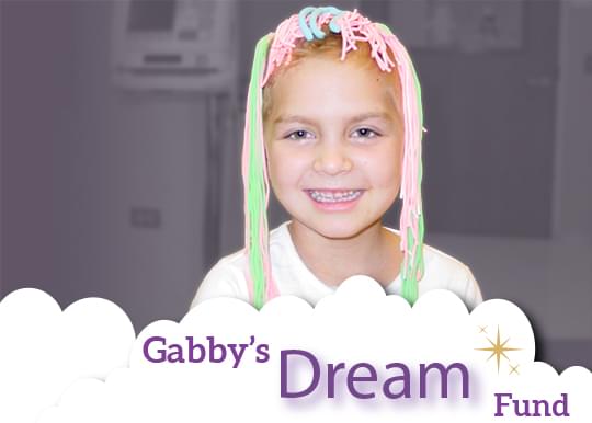 Gabby smiling at us. She has these fun and silly green, pink, and blue strings of yarn covering the top of her head and hanging down both sides of her head. She has such a genuine happy smile on her face. The background is grayed out but she is in full color and there are these illustrated clouds overlaying her at the bottom of the image. In the clouds we see the words, Gabby's Dream Fund.