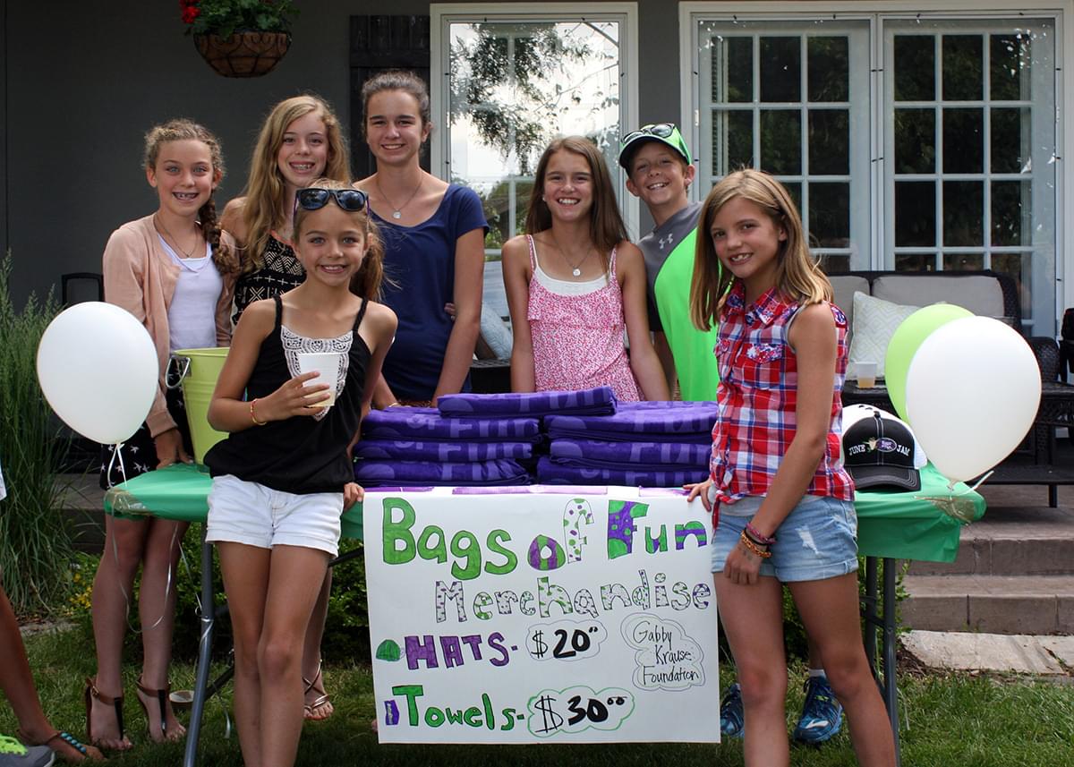 7 preteen kids standing around a table outside of a house smiling at the camera. There are Bags of Fun Towels and hats on the table that they are selling. There is a sign hanging from the front of the table that says, Bags of Fun Merchandise. Hats - $20, Towels - $30. Gabby Krause Foundation.