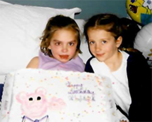Gabby and a friend sitting together posing for the camera. They are holding a large white cake with sprinkles, words that are hard to make out, and some sort of a pink character on it. Gabby appears to be in bed because there are white pillows behind her as well as a balloon. She looks like she is in the hospital.