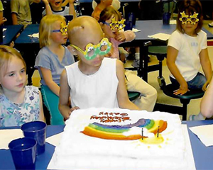 Gabby is sitting down at a blue table and looking at a large white birthday cake with a rainbow on it as well as words that are hard to make out. She is looking at the cake and has some goofy green and yellow glasses on. There are kids sitting at the table behind her and there appears to be one on either side of her, but they are mostly cut off in this photo. It appears to be a birthday party for Gabby.
