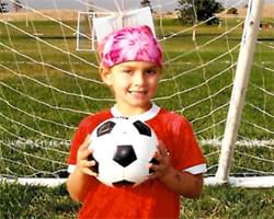 Gabby is holding a soccer ball in front of her and is smiling at the camera. She is wearing her red soccer jersey and she has a pink flower bandana on her head. We see the soccer goal net behind her and a grassy field.