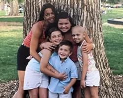 One adult and 4 kids are standing at the base of a large tree. They all have their arms around each other and are smiling. Gabby is one of the kids in the group. Behind the tree we see grass and what looks like the edge of a playground or mulch area.
