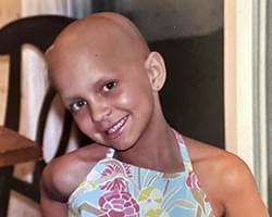 A shoulders up shot of Gabby smiling with her head slightly titled. She is wearing a pink, baby blue, and light green shirt or dress with geometric flower shapes on it. She is bald because of the cancer treatments she has had but she looks happy in this moment.