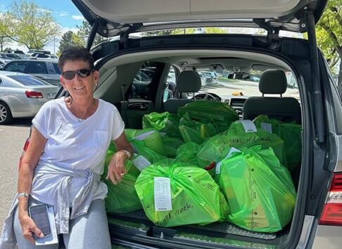 Susan sitting in the open trunk of her van with lots of green bags in the trunk. She is wearing sunglasses, a white short sleeve shirt, and a sweater tied around her waster. She appears to be in a parking lot.