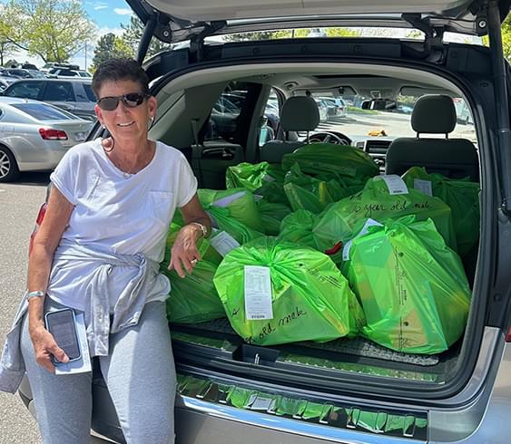 Susan sitting in the open trunk of her van with lots of green bags in the trunk. She is wearing sunglasses, a white short sleeve shirt, and a sweater tied around her waster. She appears to be in a parking lot.