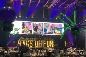 A stage with large light up yellow letters with the words Bags of Fun written out sitting on the stage. There is a large screen above the letters with images of bag recipients and there are tables in front of the stage nice set ready for guests to arrive. There are a couple people in front of one table getting ready to site down.