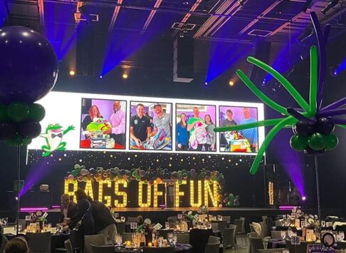 A stage with large light up yellow letters with the words Bags of Fun written out sitting on the stage. There is a large screen above the letters with images of bag recipients and there are tables in front of the stage nice set ready for guests to arrive. There are a couple people in front of one table getting ready to site down.