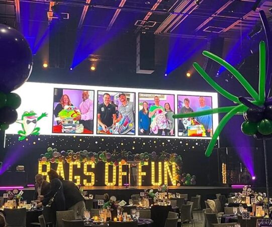 A stage with large light up yellow letters with the words Bags of Fun written out sitting on the stage. There is a large screen above the letters with images of bag recipients and there are tables in front of the stage nice set ready for guests to arrive. There are a couple people in front of one table getting ready to site down.