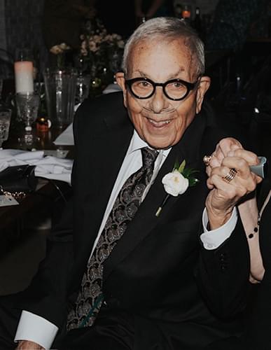 Mike Solomon in a dark suite and tie with a flower on his lapel He is holding someones hand and dancing but we only see a close up of him and the hand. He is smiling and has dark rimmed glasses on.