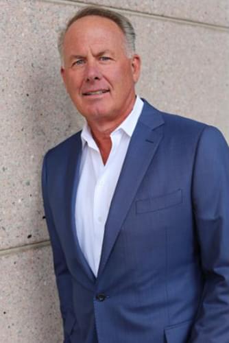 Sandy Hamilton in a blue shirt and open white collared shirt. he is leaning against a wall.