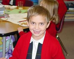 Gabby with short hair smiling with her open mouth smile at us. She is wearing a red cardigan style sweater with a white and black collared shirt underneath. She appears to be in a classroom and she looks very happy.