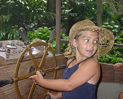 Gabby with a straw hat on sitting in some sort of wooden fake vehicle with a spinning steering wheel. She is look back and there is lots of green tropical trees behind her.