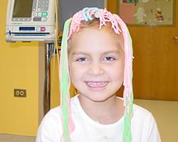 Gabby smiling at us. She has these fun and silly green, pink, and light blue strings of yarn covering the top of her head and hanging down both sides of her head. She has such a genuine happy smile on her face. Sh is in a hospital or doctors office. We can see a medical monitor behind her.