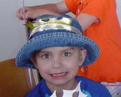 A close up of Gabby smiling at the camera. She is wearing a blue circle rimmed hat with something shinny on the front and a blue shirt.