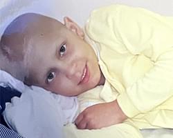 A close up of Gabby lying down on a white pillow and smiling at us. She is wearing what appears to be yellow pajamas.