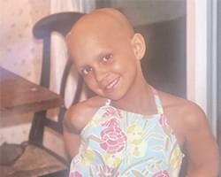 Gabby smiling and wearing a blue summer dress with pink and yellow flowers. She does not have any hair, which suggests this is after her chemo treatments had started. She does appear to be really happy though.