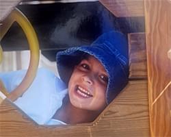 Gabby is peering through a hole in some sort of a wooden structure. She has an open smile on her face and she is wearing a blue bucket hat.