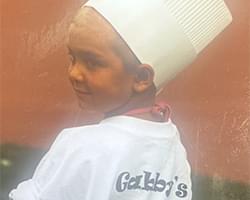 Gabby's back is turned to us and she is looking over her shoulder. She has a white chefs hat on and she is wearing a white shirt with the word Gabby's visible on it. It looks like there is a word under it but it is cut off.