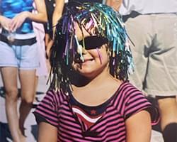 Gabby with black sunglasses on and a silly purple, blue, and green confetti wig on. She has a slight smile on her face and she is wearing a pink and black striped shirt.