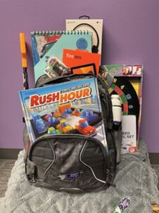 A Bags of Fun backpack with Rush Hour traffic jam logic game in the front and a Bags of Fun water bottle in the side cup holder, and a Amazon Fire, art pad and headphones in the back. The Bags of Fun backpack is sitting on a Bags if Fun blanket, and it is resting on a packaged dart board.