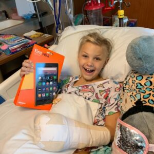 Kid in his hospital bed holding an Amazon Kindle and smiling. He has a large cast on his arm and a stuffed animal next to him.