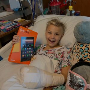 Kid in his hospital bed holding an Amazon Kindle and smiling. He has a large cast on his arm and a stuffed animal next to him.